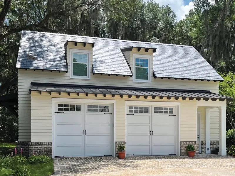 Residential garage with two white garage doors, featuring decorative windows and a stylish roof, surrounded by greenery, illustrating Overhead Door Company of The Desert™'s durable garage door offerings.