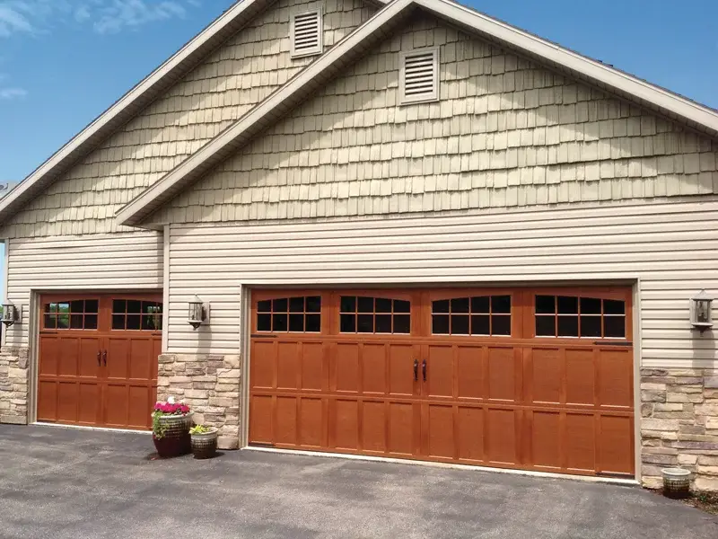 Garage with two wooden carriage-style doors featuring windows, stone accents, and a light fixture, set against a blue sky, representing residential garage door installation services by Overhead Door Company of The Antelope Valley™.