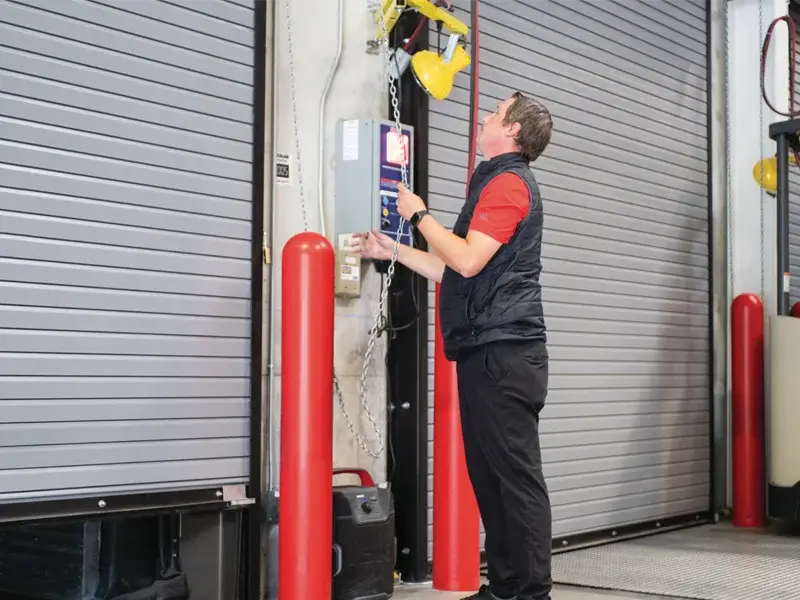 Technician operating a control panel for a heavy-duty rolling steel service door in a commercial setting, emphasizing door installation and maintenance services.