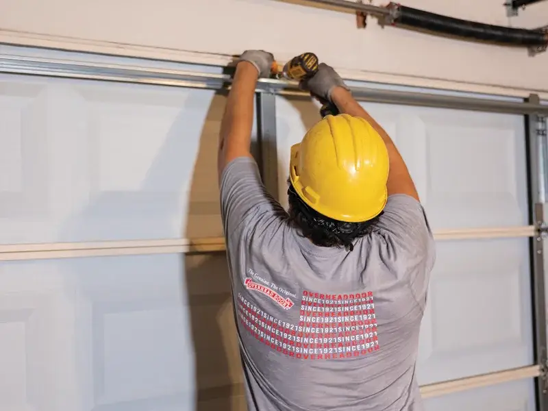 Technician installing a garage door track, wearing a yellow hard hat and gray shirt with Overhead Door branding, demonstrating garage door replacement services.