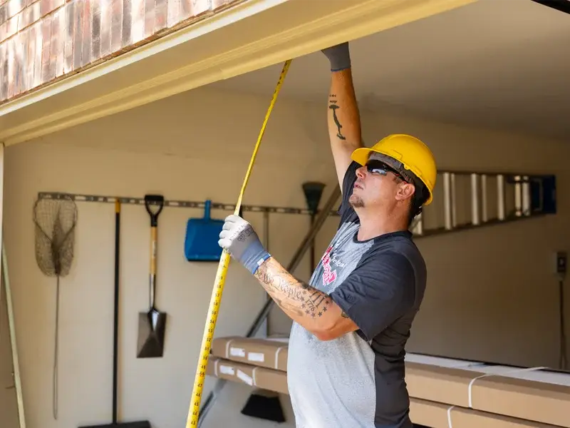 Worker measuring garage door opening for installation, wearing safety gear, with tools and materials visible in the background, emphasizing professional garage door services by Overhead Door Company of the Antelope Valley™.