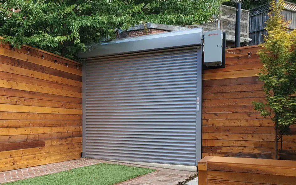 Modern silver roll-up garage door in a residential setting with wooden fence and greenery, showcasing stylish and secure design for Coachella Valley homes.